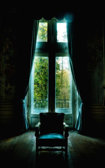 An empty upholstered chair facing tall ruined windows in a derelict room, daylight and overgrowth beyond.